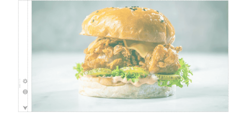 Hamburguesa con pollo frito, lechuga, rodajas de pepinillo, queso y una salsa, servida en un pan suave y dorado. La hamburguesa está presentada sobre una superficie blanca y tiene un fondo neutro, resaltando los ingredientes frescos y el crujiente del pollo.