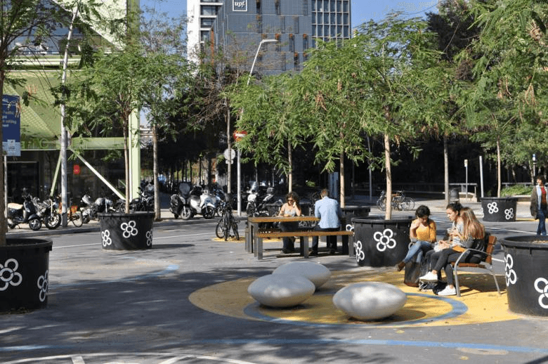 Espacio urbano (supermanzana) en Barcelona, al aire libre. Con árboles jóvenes en maceteros grandes negros decorados con flores blancas. Hay varias personas sentadas en bancos y en una mesa, socializando o usando sus dispositivos móviles. En el centro, un área circular amarilla y azul contiene varias estructuras de piedra redondeadas para sentarse. Al fondo, hay edificios modernos y un aparcamiento de bicicletas.