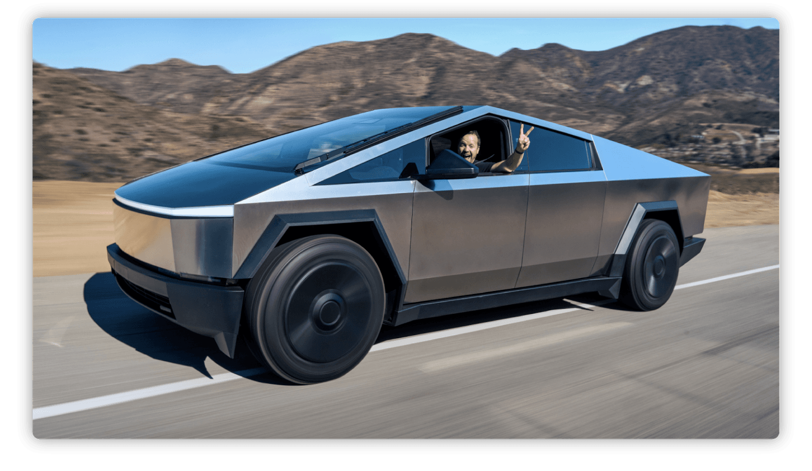 Imagen de una camioneta futurista de diseño angular y metálico, identificable como el Cybertruck de Tesla, circulando en una ruta. En el asiento del conductor, una persona sonriente saca la mano por la ventana y hace el signo de paz. Al fondo se observan montañas desérticas bajo un cielo despejado.