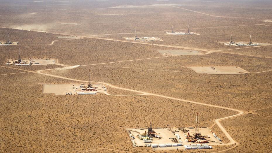 Fotografía aérea de un área de explotación petrolera en una zona árida y extensa, se observan múltiples plataformas de perforación distribuidas en la tierra, conectadas por caminos de tierra que serpentean entre las instalaciones, el paisaje está mayormente despoblado y cubierto de vegetación baja típica de zonas desérticas, la imagen destaca la extensión de la operación y el impacto en el entorno natural, representando un espacio dedicado a la extracción de recursos en una región remota.